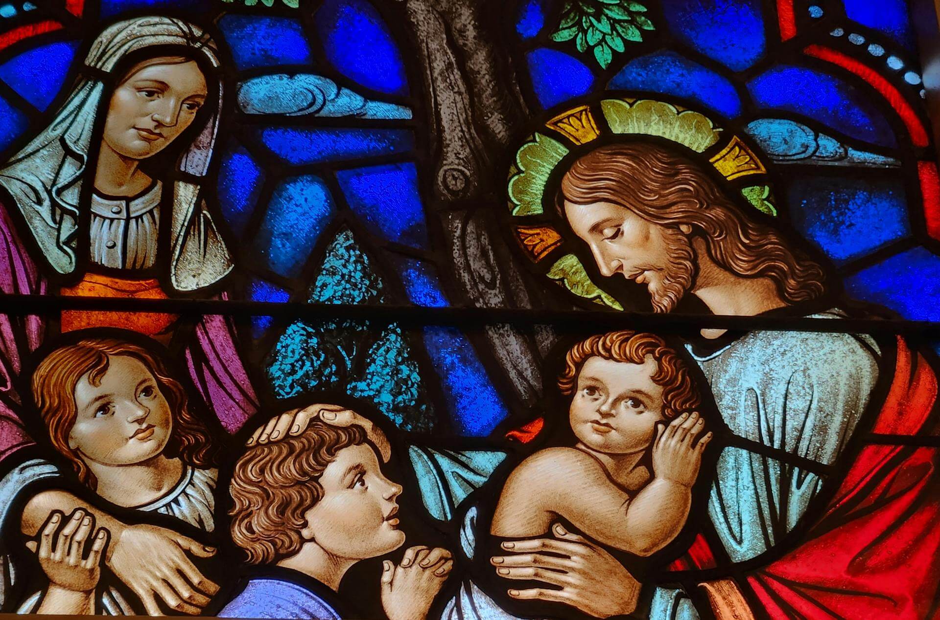 Colored stained glass depiction of Jesus with a halo surrounded by children and a woman in biblical attire under a tree, emphasizing themes of care and surrender.