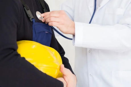 A physician listens to the heartbeat of a worker holding his yellow hard hat.
