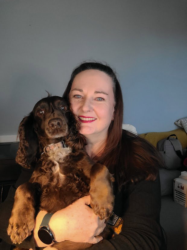 Smiling site owner hugging her brown cocker spaniel on her lap, both looking directly into the camera