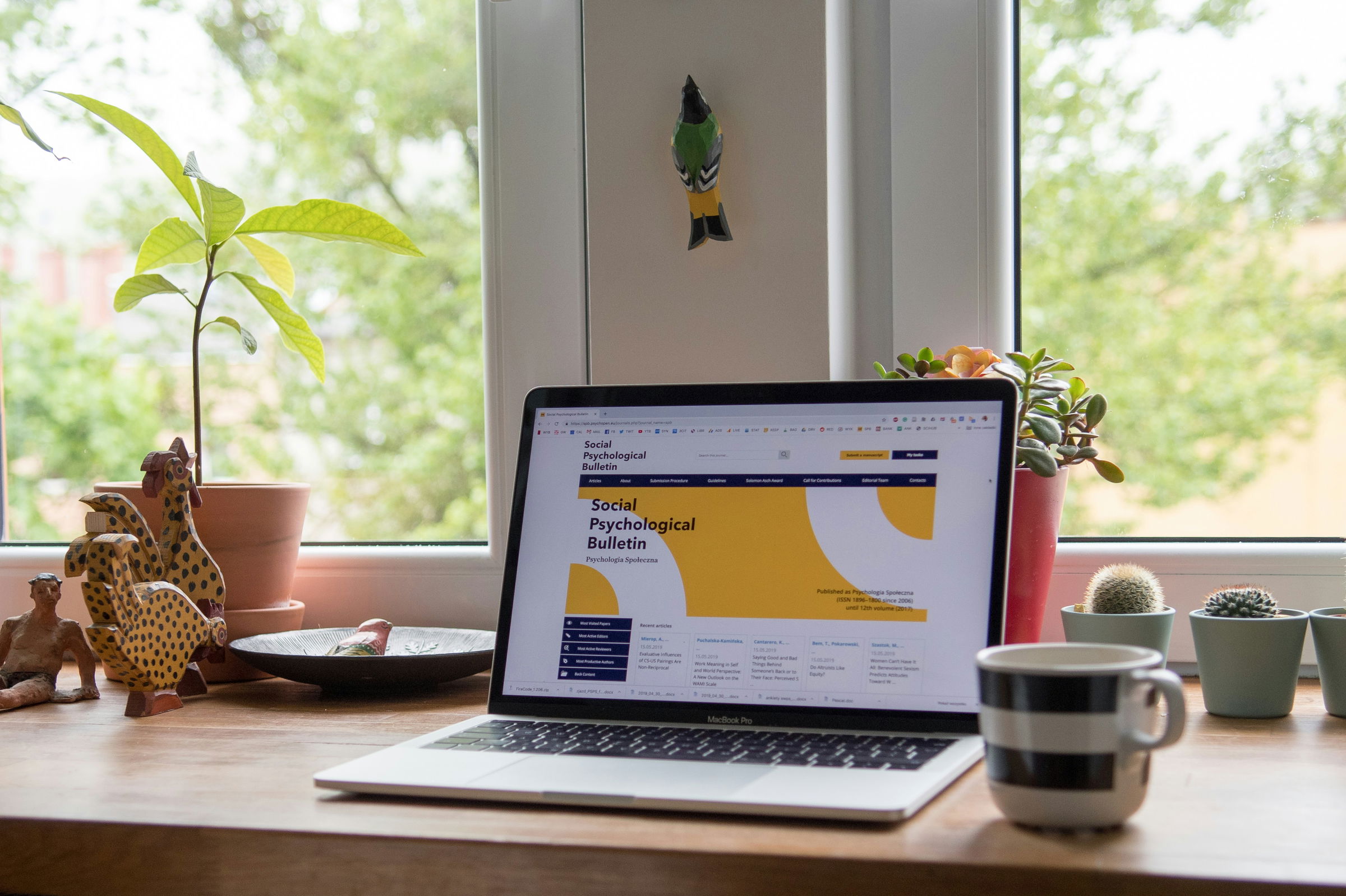 A laptop on a wooden desk shows an academic journal site, surrounded by plants, a mug, and decorative items by a window.