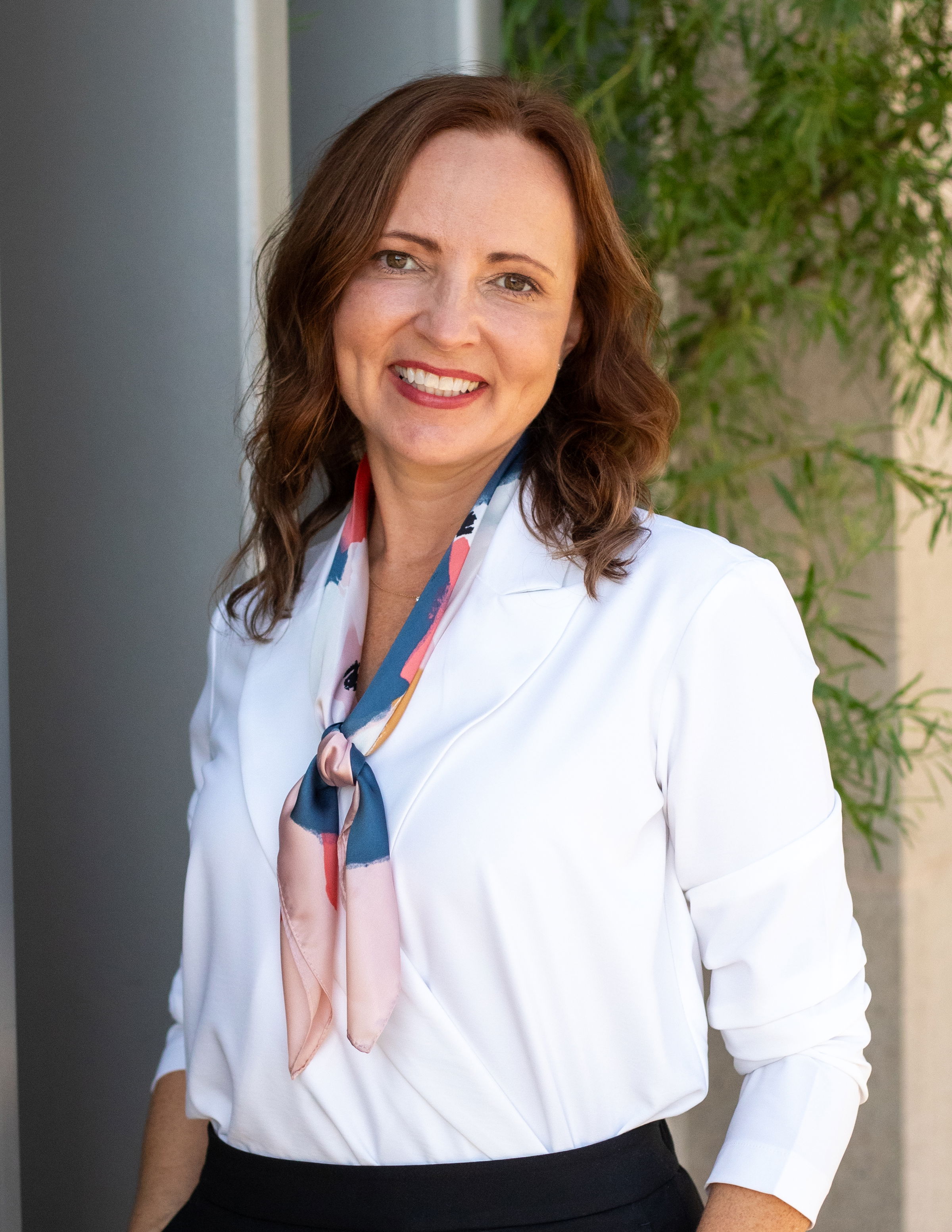 Professional headshot of Carrie Moss, smiling in a white blouse with a patterned scarf, standing outdoors.